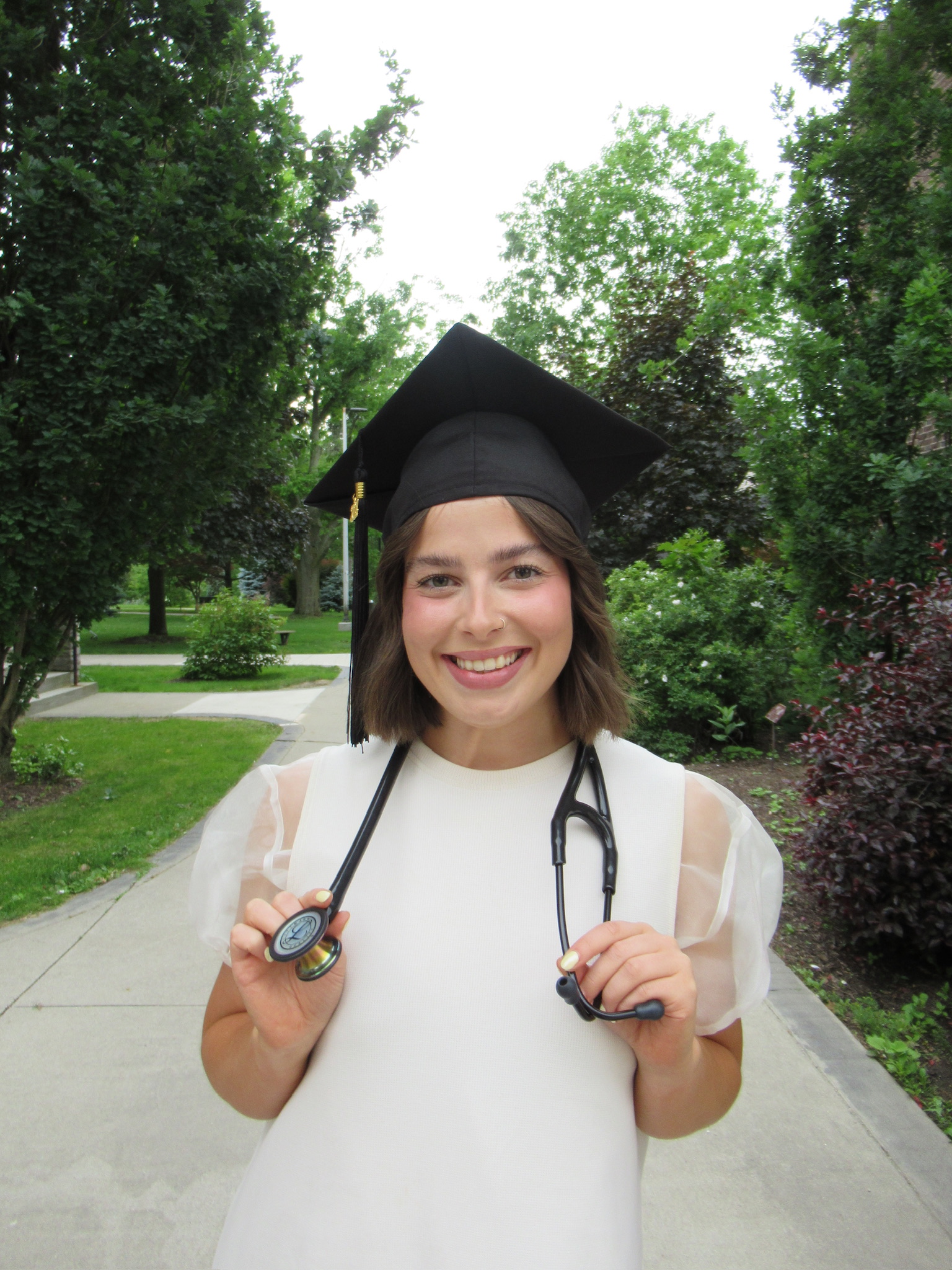 Woman outside wearing a black grad cap. She has short brown hair with a black stethoscope on her neck. She is wearing a white blouse. 