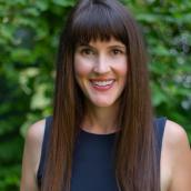 A woman with long brown hair with bangs is standing in front of a green leafy bush wearing a black tank top and smiling into the camera.