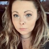A woman with wavy blonde hair looks at the camera, she has a necklace and black shirt on.