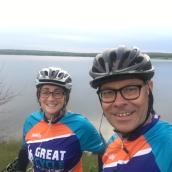 A woman wears glasses, a bike helmet and bike racing shirt, as does the man standing next to her. They are smiling at the camera, taking a selfie.