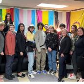A group of 13 people stand together in front of a large LGBTQ+ flag.