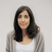 A woman with long brown hair and eyes is wearing a white, scoop-neck shirt with a beige pullover sweater, smiling at the camera.