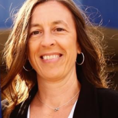 A woman with long brown hair and eyes, hoop earrings wears a white shirt with a black sweater and is smiling at the camera.