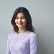 A woman with medium-length brown hair wears a light purple sweater and a simple necklace and is smiling at the camera.