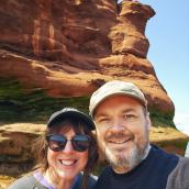 Woman with a hat and sunglasses wearing a black sweatshirt is standing beside a man with a beard and moustache wearing a hat and black sweatshirt. They are standing in front of a tall rock formation outdoors, smiling at the camera.