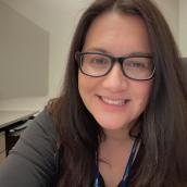A woman with long, brown hair, glasses, and a black long-sleeve shirt sits in an office room and is smiling at the camera.