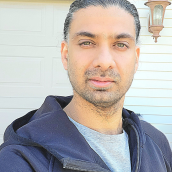 A photo of a man with black hair and beard, wearing a black hoodie over a grey shirt. He is standing in front of a house.