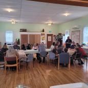 A group of people is gathered around a table in an indoor setting. The atmosphere suggests a living or dining room environment. 