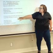 A woman is pointing at a projector screen during a presentation. The content appears to focus on reaching out for support, featuring contact information for various mental health and youth services in Canada. The setting is indoors, with the woman dressed casually in jeans and trousers, engaged in sharing important resources.