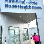 A woman is standing in front of the Dr. Maria Angwin Memorial Wyse Road Health Clinic. The setting is outdoors, featuring a building with a sign that identifies it as the clinic.