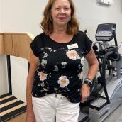 A woman with light, brown hair wears a black shirt, white pants and is standing, smiling at the camera.