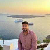 Arjun Sethi, wearing a light red button-up shirt and glasses, sits on a white terrace overlooking the ocean at sunset. Behind him, the calm water reflects the warm glow of the sky, with two cruise ships anchored near a rocky island in the distance.