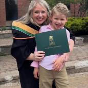 Keshia MacDonald, with long, blond hair, is smiling with her son, also with blond hair, while wearing a graduation gown and holding a diploma.
