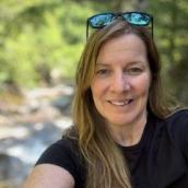 Woman with long, blonde hair is wearing a black t-shirt and sunglasses on her head and is smiling at the camera.