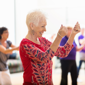 Woman with short, white hair, earrings, and a red shirt with black embellishments is snapping her fingers in a room with other people blurred in the background.