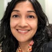 A woman is smiling for the camera. She has a joyful expression. The portrait captures her indoors; a typical look consistent with portrait photography. 