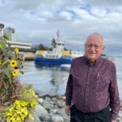 A senior man with gray hair and glasses is wearing a dark purple shirt while standing outside in front of the ocean, rocks and a ferry is in the background.