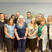 A group of 13 people in casual business attire stand together and smile at the camera.