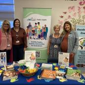 A group of four women stand side-by-side behind a table with a colourful display of promotional materials and smiling at the camera