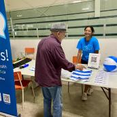 : Roslyn George, Northern Zone Health Ambassador, standing behind an information table with pamphlets, speaking with a community member at a local event.