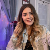 A woman with long, blonde hair is in a bedroom wearing a jean jacket and smiling at the camera.