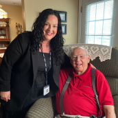 A woman with curly, brown hair and a black shirt and sweater with beige pants sits beside a senior man with a red shirt and suspenders in a living room.