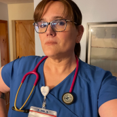 A photo of a woman with brown hair in a ponytail, she is wearing black framed glasses, blue scrubs with a red stethoscope around her neck, and a badge clipped to her collar. 