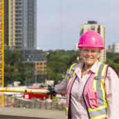 Woman wearing a pink hard hat and safety vest stands with a view of a construction site behind her, smiling at the camera.