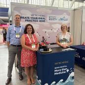 Three Nova Scotia Health representatives stand at a recruitment booth featuring a banner that reads “We’re hiring — Create your ideal practice here. Nova Scotia: More than Medicine.” The group is smiling, and the display table is covered with promotional materials.