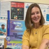A woman with a dark blonde bob wears a mustard yellow shirt and a pendant necklace while smiling at the camera in front of a bulletin board with colourful posters on it.