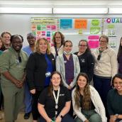 A group of 13 healthcare workers stand together in front of a brightly lit whiteboard, smiling at the camera.