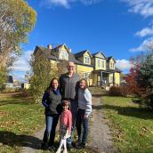 A photo of a family of four in front of a yellow home. The woman on the left is wearing a blue sweater with a black vest on top and blue jeans. She has brown curly hair. Man in the back is bald with glasses wearing a black shirt and blue jeans with a brown belt, girl on the right has curly brown hair with a light blue sweater and blue jeans. Girl in the front has brown short hair with a pink headband on, she is wearing a pink sweater with a plaid pink, black and white skirt and white tights. 