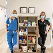 Two women wearing doctor scrubs stand beside a bookshelf full of food and personal care products.