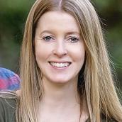 A woman with long, blonde hair stands outdoors and is wearing a green shirt.