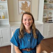 A woman with long brown hair wears a blue dress with a brown belt and is sitting in a room with white cabinets behind her.