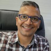 A person with short hair and glasses is smiling while seated in an office chair, wearing a plaid shirt.