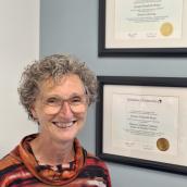 Lenora Brace, nurse practitioner, smiling while standing in front of two framed Athabasca University diplomas on a light blue wall and wearing a striped orange and brown sweater.