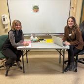 Alice Murphy (left) and Natasha Osmond (right), two Cape Breton diabetes educators, sit at a table in a clinic room. Alice is writing on a clipboard, and Natasha is working on a laptop. Both are smiling toward the camera, with educational materials and medical supplies on the table and a whiteboard behind them.