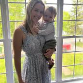 A woman with blonde hair stands in front of a window wearing a white dress with a floral print, holding a baby, and is smiling at the camera.