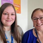 Two women sit side-by-side, one has long blonde hair and the other has brown hair in a ponytail and glasses.