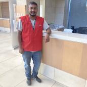 A man with a red best and jeans stands at a front desk in the lobby of a healthcare centre, smiling at the camera.