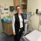 A person wearing glasses, a white long-sleeve shirt, and a black vest is standing in a medical examination room beside medical equipment and supplies.