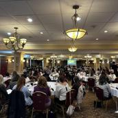 Groups of people sit at tables in a large conference room.