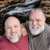Two men standing in front of a waterfall with a fall leaves. Man on the left has a gray beard, wearing a red sweater. Man on the right has gray hair and gray beard, wearing a brown/grey sweater. 