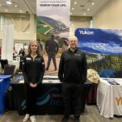 Woman standing on the left, man on the right. Both wearing black and they are standing in front of information tables.