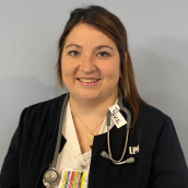 A healthcare worker wearing a black jacket with “LPN” on it smiles at the camera. She has a stethoscope around her neck and stands in front of a plain light-coloured background.  