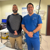 Two men standing together smiling. One in casual clothes and the other in blue scrubs. They are in a hospital room.