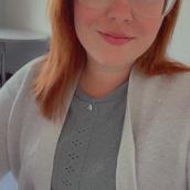  Photo of Shannon Goucher, woman with shoulder-length red hair and clear-framed glasses smiling at the camera, wearing a light grey knit top and cardigan, seated indoors.