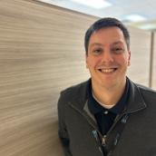 A man with short brown hair stands against a wood-panel wall wearing a zip-up sweater and a golf shirt, and is smiling at the camera.