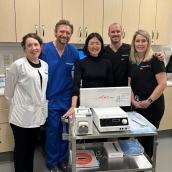 Five people stand together in front of a piece of hospital equipment smiling at the camera.